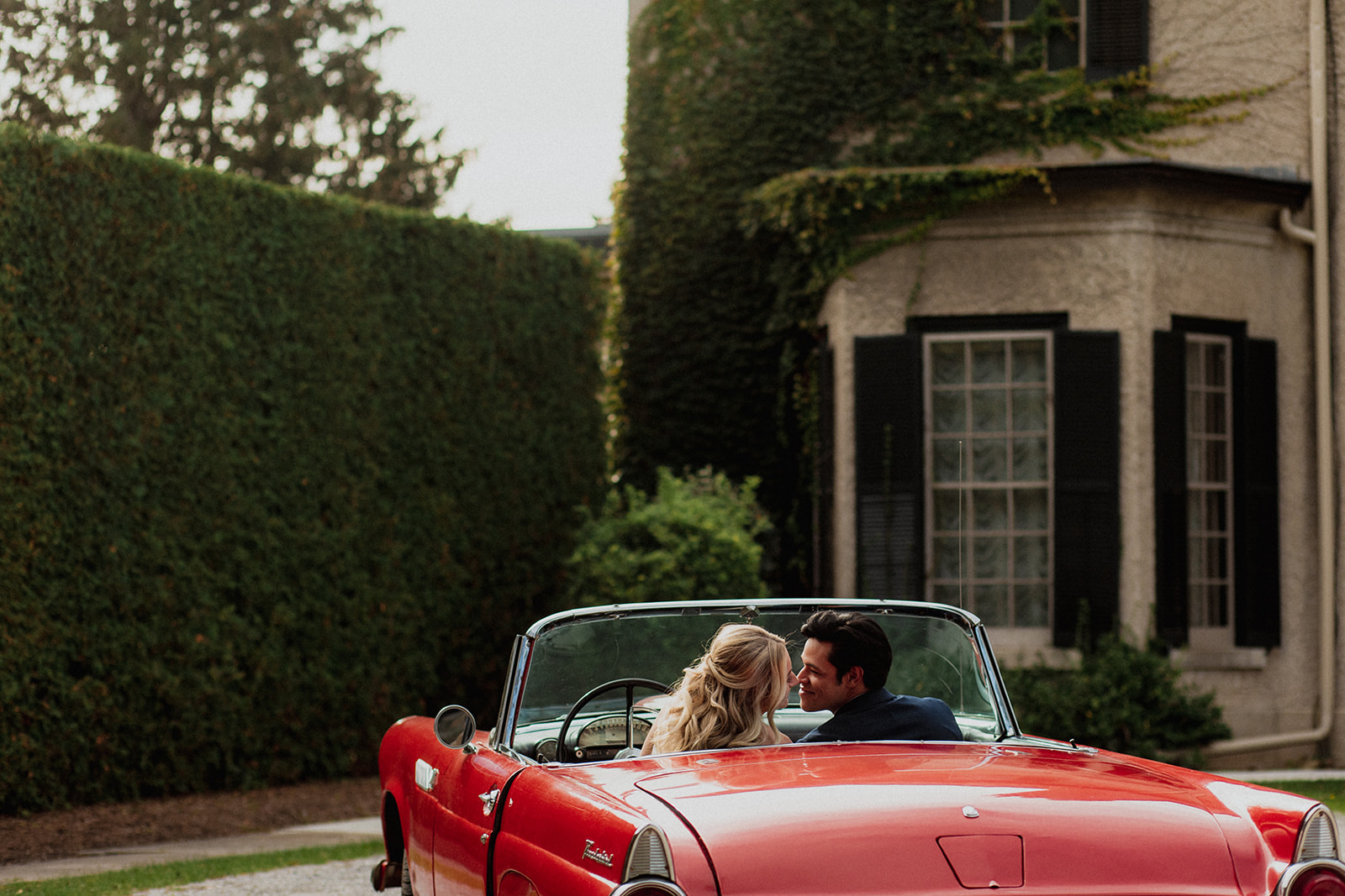 A couple shares a moment in a classic red convertible parked beside a lush green hedge near a stately building.